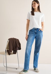 Femme debout, une main dans la poche, portant une chemise blanche à manches courtes, un jean bleu et des baskets, à côté d'une chaise sur laquelle est posée une veste marron.