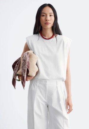 Femme aux longs cheveux noirs portant un haut blanc sans manches et un pantalon, collier de perles rouge, tenant un sac tissé avec une écharpe marron à motifs.