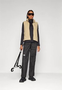 Γυναίκα που φοράει γυαλία ηλίου, μπεζ γιλέκο φλις, μαύρη μπλούζα με μακρύ μανίκι, μαύρο παντελόνι και μαύρες μπότες, κρατώντας σκιέ poles ενάντια σε απλό φόντο.