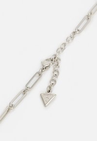 Collier chaîne en argent avec des maillons rectangulaires et un petit pendentif logo Guess triangulaire près du fermoir sur un fond blanc.
