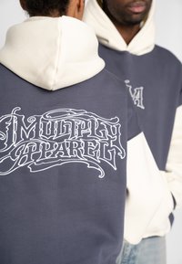 Het capuchontrui heeft een donkergrijze body met crème accenten. Het ontwerp aan de achterkant bevat "Multiply Apparel" in witte, ingewikkelde letters.