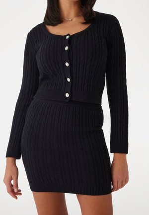 Femme portant un pull court à boutons en maille torsadée noire et une mini-jupe assortie en maille torsadée noire sur fond blanc.