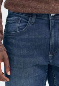 Temno modri kavbojke iz denima s klasičnim pet-pocket dizajnom, z rahlim zbledelostjo in kontrastnim šivanjem. Tkanina ima gladek, teksturiran zaključek.