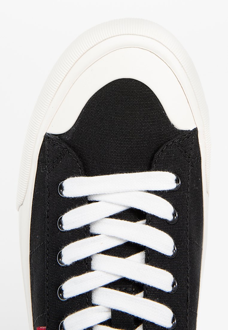 Zapatilla de lona negra con suela de goma blanca, con cordones blancos y punta redondeada. Parte superior texturizada con un patrón de costura.