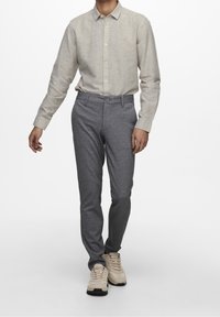 Uomo che indossa una camicia beige con bottoni, pantaloni grigi slim-fit e sneakers beige, in piedi contro uno sfondo bianco.