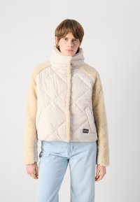 Geacă puffer cu un corp bej matlasat și mâneci pufoase de culoare nisipie. Guler înalt cu fermoar frontal. Mic logo negru în partea stângă jos.