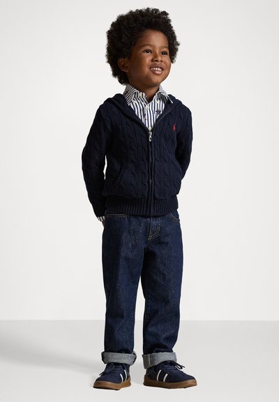 Μπλε καλσόν με πλέξη, με φερμουάρ, συνδυασμένο με καρό πουκάμισο, τζιν παντελόνι και μπλε αθλητικά παπούτσια με λευκές ρίγες. Ομαλή, ευπροσάρμοστη εμφάνιση.