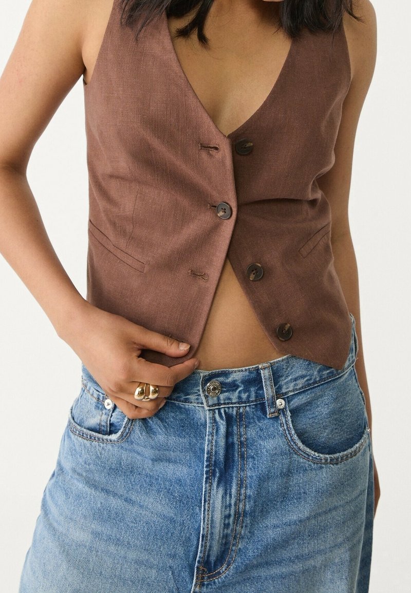 Personne portant un gilet marron sans manches boutonné et un jean en denim bleu taille haute, tenant l'ourlet du gilet d'une main.