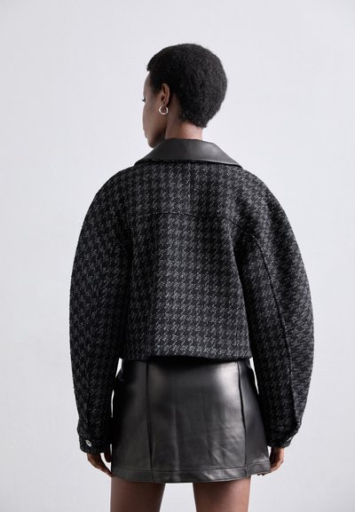 Zrezaná bunda v čierno-sivom vzore houndstooth, so čiernym koženým golierom a dlhými, nadýchanými rukávmi. Model má na sebe čiernu koženú sukňu.