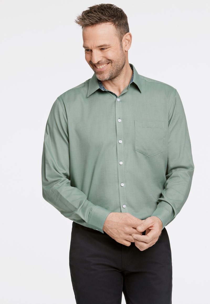 Camicia button-up verde chiaro con un tessuto testo, maniche lunghe e taschino sul petto. Presenta un colletto a fantasia. Indossata con pantaloni neri.