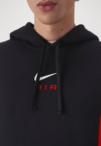 Sweat-shirt noir avec poche kangourou, arborant un logo Nike blanc et le texte "AIR" rouge. Tissu doux avec cordons de serrage et poignets côtelés.