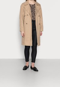 Beige dvouřadový trenčkot přes černý vzorovaný top, spárovaný s černými legínami a mokasíny se zlatými akcenty.