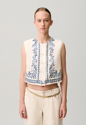 Joven mujer que lleva un top corto sin mangas color crema con bordado floral azul y pantalones crema de cintura alta con cinturón de cadena dorada.