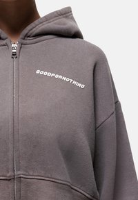 Felpa con zip in un tessuto grigio tenue, caratterizzata da una vestibilità comoda, chiusura frontale a zip e testo "GOODFOR NOTHING" in bianco sul petto.