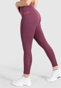 Burgundy korkeavyötäröiset leggingsit sauman yksityiskohdilla ja teksturoitujen paneelien kanssa, joissa on pieni logo takana ja ne on yhdistetty valkoisiin tennareihin.