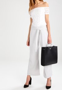 Femme portant un haut en dentelle blanche à épaules dénudées et un pantalon large blanc, tenant un sac à main noir avec des poignées métalliques rondes, portant des talons noirs.