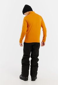 Pomarańczowa bluza z długim rękawem, wysoka kołnierz i czarne wodoodporne spodnie. Na głowie czarna dziana czapka. Widoczne czarne obuwie. Prosty, funkcjonalny design.