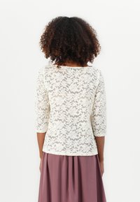 Femme aux cheveux bouclés portant un haut en dentelle florale blanche à manches trois-quarts et une jupe plissée mauve, de dos.