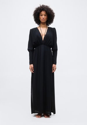 Mujer con cabello rizado, vestida con un vestido largo negro con escote en V profundo y mangas largas, de pie descalza contra un fondo liso.