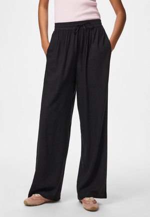 Άτομο που φοράει φαρδιά μαύρη παντελόνα με κορδόνι, ανοιχτό ροζ αμάνικο τοπ και μπεζ casual παπούτσια, στέκεται με τα χέρια στις τσέπες.