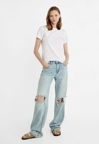 T-shirt blanc en coton associé à un jean délavé bleu clair avec de grandes déchirures au niveau des genoux et une coupe décontractée. Des sandales avec une lanière marron complètent le look.