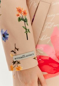 Bežová tkanina s farebnými kvetinovými vzormi v oranžovej, ružovej a modrej. Obsahuje štítok s nápisom "THE BUTTERFLY EFFECT" v čiernom píme.