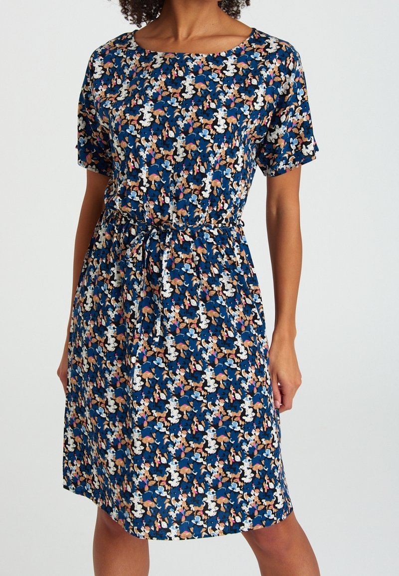 Femme portant une robe bleue mi-genoux avec manches courtes, taille cintrée et motif floral abstrait multicolore.