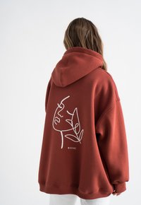 Übergroßer rostfarbener Hoodie mit Kordelzugkapuze. Verfügt über ein weißes abstraktes Gesichtsmotiv und den Schriftzug "MIRONS" auf dem Rücken. Weicher, strukturiertes Material.