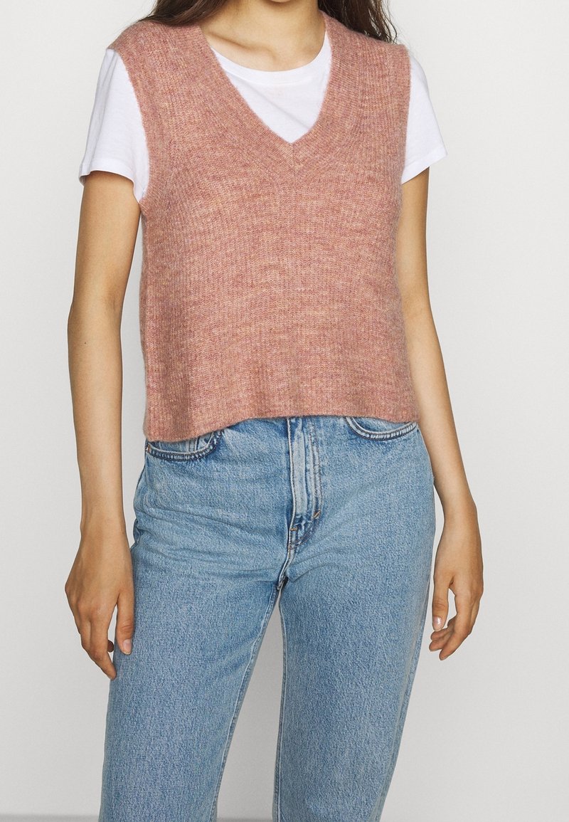 Frau in einem weißen kurzärmligen Hemd unter einem pinkfarbenen gestrickten ärmellosen Pulloverweste, kombiniert mit hochgeschnittenen blauen Jeans.