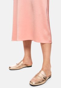 Goldfarbene, metallisch schimmernde Slipper mit runder Spitze und flachem Absatz, kombiniert mit einem rosafarbenen, seidigen Kleid, das bis zur Mitte der Wade reicht.