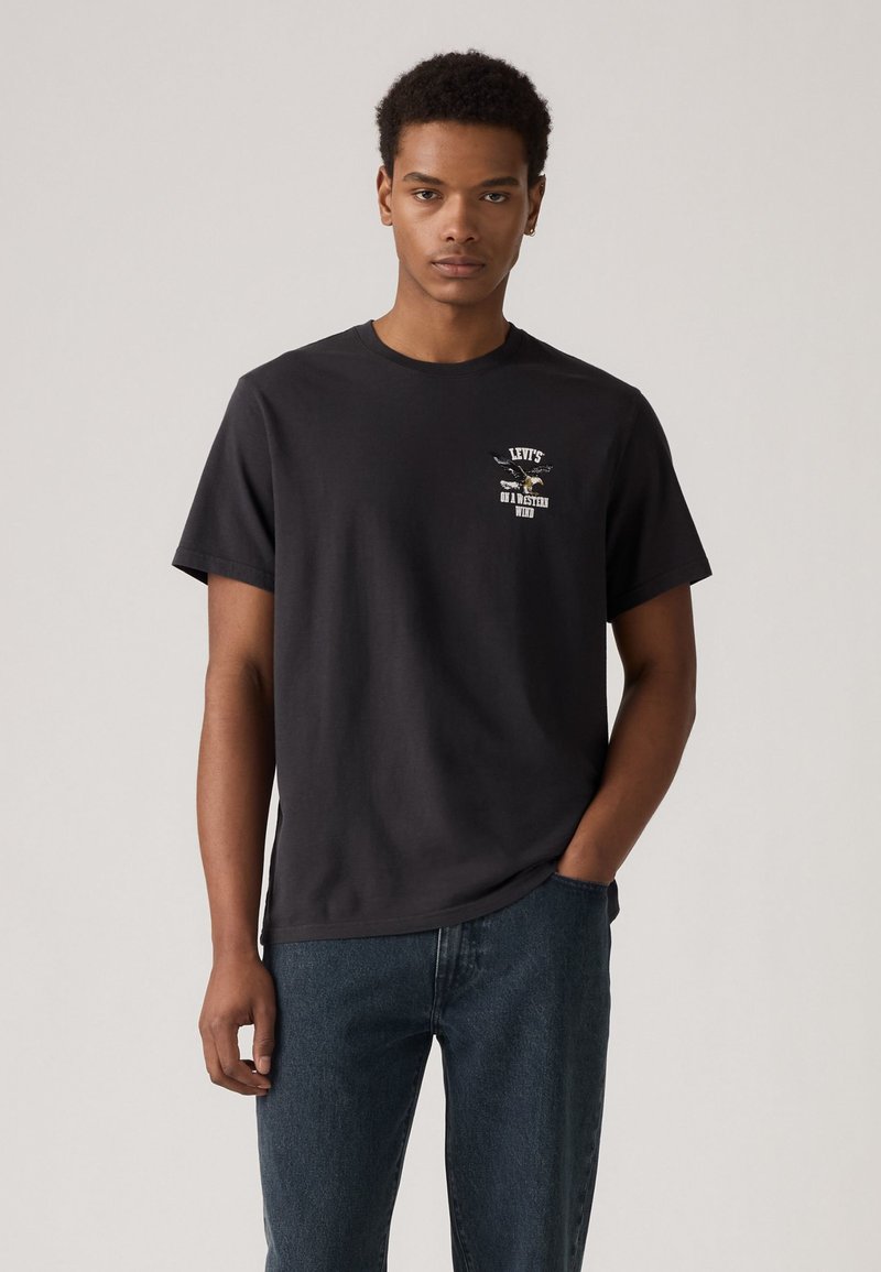T-shirt en coton noir à manches courtes, avec un imprimé graphique et un texte sur la poitrine ; associé à un jean en denim foncé.