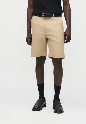 Mand iført beige knælange shorts med sort bælte, sorte sokker, sorte loafers og en sort skjorte stoppet ind, stående mod en hvid baggrund.