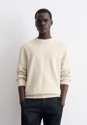 Beige knitted sweater with ribbed crew neck and cuffs. Features a subtle logo on the chest. Paired with dark trousers.