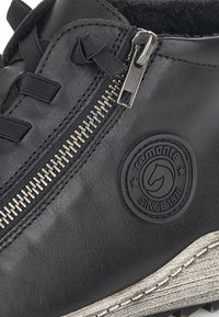 Chaussure en cuir noir avec fermeture éclair sur le côté, patch logo texturé et coutures bien visibles. Semelle en caoutchouc avec une accentuation blanche.
