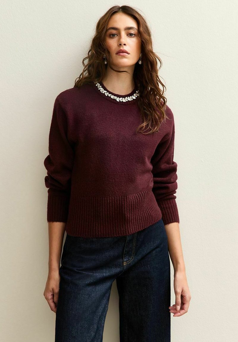 Maglione lavorato a maglia color bordeaux con orlo e polsini a coste, caratterizzato da un collo decorativo con perline. Abbinato a jeans a gamba larga in denim scuro.