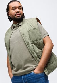 Άνδρας με πλεξούδες στα μαλλιά, φορώντας ελιάς πράσινο γιλέκο με επένδυση και πλεκτό πουκάμισο, με τα χέρια στις τσέπες μπλε τζιν, κοιτάζει πλάγια μπροστά σε λευκό φόντο.