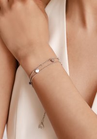Silver layered bracelet featuring delicate chains, circular and oval charms, with a textured finish and a small leaf-shaped accent at the end.