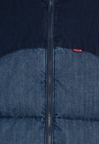 Mörkblå kordflod och denim väst med en central dragkedja, vadderad struktur på den nedre delen och en liten röd Levi's logotags.