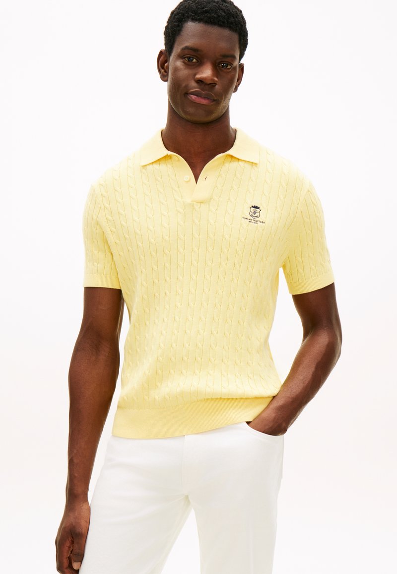 Άνδρας που φοράει κίτρινο πλεκτό πόλο με κοντά μανίκια και λευκό παντελόνι, στέκεται με το ένα χέρι στην τσέπη σε λευκό φόντο.