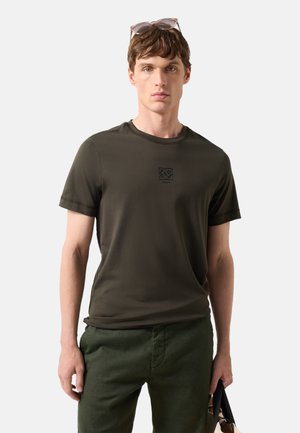 Ung mand iført mørkegrøn t-shirt og bukser, solbriller på hovedet, holder en tofarvet taske med sorte håndtag.