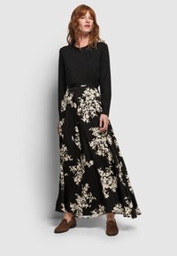 Frau, die ein schwarzes Langarmoberteil, einen schwarz-weißen floral gemusterten Maxikleid und braune Slipper trägt, steht vor einem schlichten hellen Hintergrund.