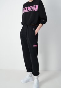 Svart sweatshir med "CHAMPION" i rosa bokstäver, ihop med svarta joggers med en rosa logga och vita sneakers. Mjuk tygkvalitet, avslappnad passform.