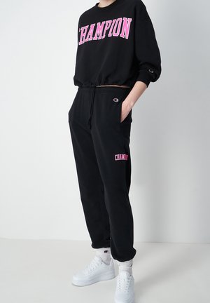 Svart sweatshir med "CHAMPION" i rosa bokstäver, ihop med svarta joggers med en rosa logga och vita sneakers. Mjuk tygkvalitet, avslappnad passform.