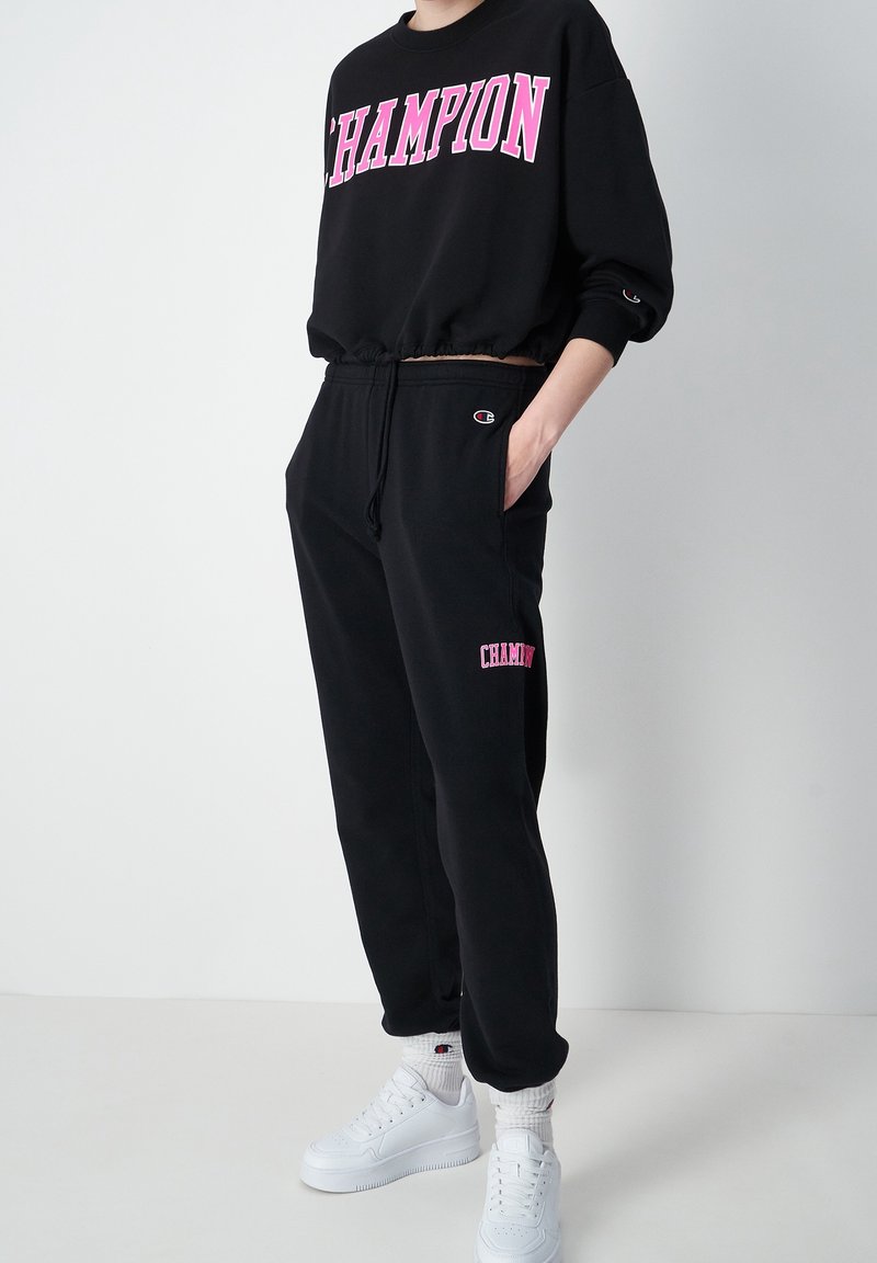 Svart sweatshir med "CHAMPION" i rosa bokstäver, ihop med svarta joggers med en rosa logga och vita sneakers. Mjuk tygkvalitet, avslappnad passform.