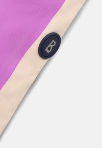 Detailní záběr na růžovou a béžovou látku s černým kulatým knoflíkem, na kterém je stříbrné písmeno "B", na čistě bílém pozadí.