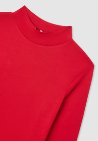 Camicetta rossa a maniche lunghe con colletto alto su uno sfondo grigio chiaro, che mostra la parte superiore del torso e la cucitura della spalla.