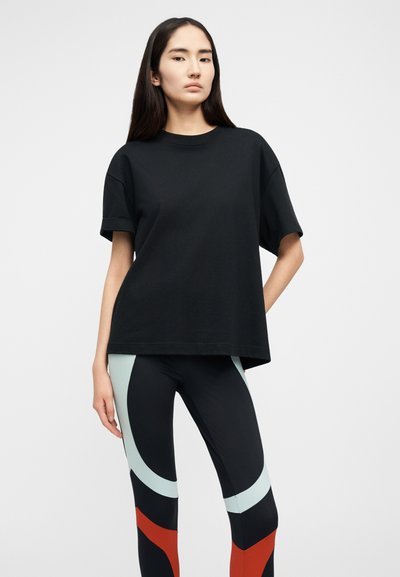 Γυναίκα που φοράει φαρδιά μαύρη κοντομάνικη μπλούζα και μαύρα κολάν με ανοιχτό μπλε και κόκκινα γεωμετρικά σχέδια, στέκεται μπροστά σε απλό φόντο.