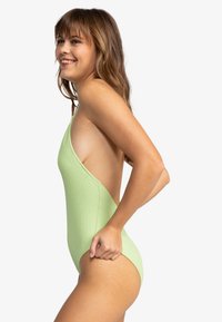 Maillot de bain une pièce vert côtelé avec une découpe profonde sur le côté, des bretelles fines et une texture lisse. Présente des jambes à découpe haute et une couverture minimale.
