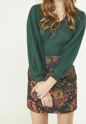 Femme portant un haut à manches longues vert foncé avec des manches texturées et une jupe à motif floral, mains jointes devant.