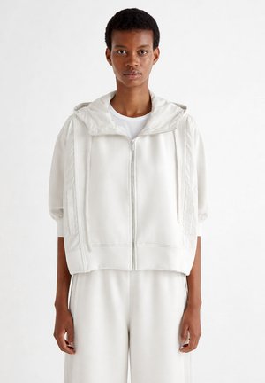 Młoda osoba nosząca luźną, białą bluzę z kapturem na suwak i pasujące szerokie spodnie, stojąca na jednolitym białym tle.